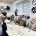 Zdjęcie przedstawia seniorów, którzy siedzą w sali w budynku DDPS+