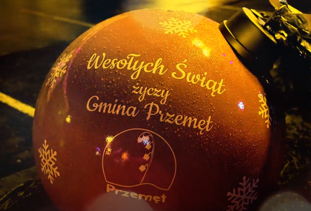 Życzenia świąteczne dla mieszkańców gminy Przemęt. Zdjęcie przedstawia czerwoną dużą bombkę z napisem "Wesołych Świąt życzy Gmina Przemęt". Na bombce znajdują się również logo Gminy Przemęt oraz białe śnieżynki.