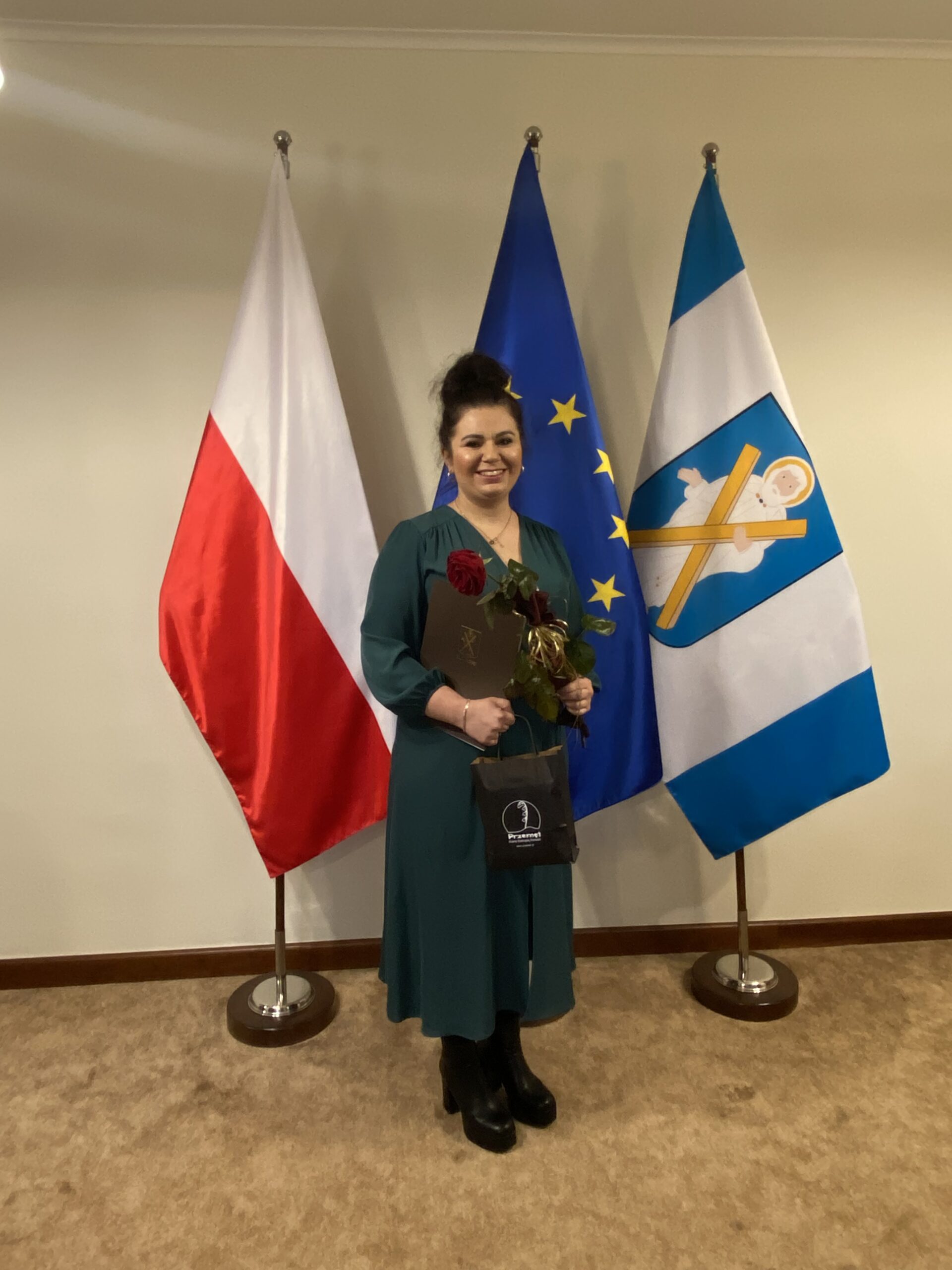 Zdjęcie przedstawia Panią Ewę Pelec, która trzyma w rękach różę oraz akt nadania. Zdjęcie wykonano w biurze wójta.
