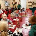 Zdjęcie przedstawia dzieci siedzące przy dużym czerwonym stole i piszą list do Świętego Mikołaja. W tle osoba przebrana za elfa.