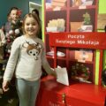 Zdjęcie przedstawia uśmiechniętą dziewczynkę, która wrzuca swój list do skrzynki z pocztą do św. Mikołaja. W kolejce czeka reszta dzieci na swoją kolej.