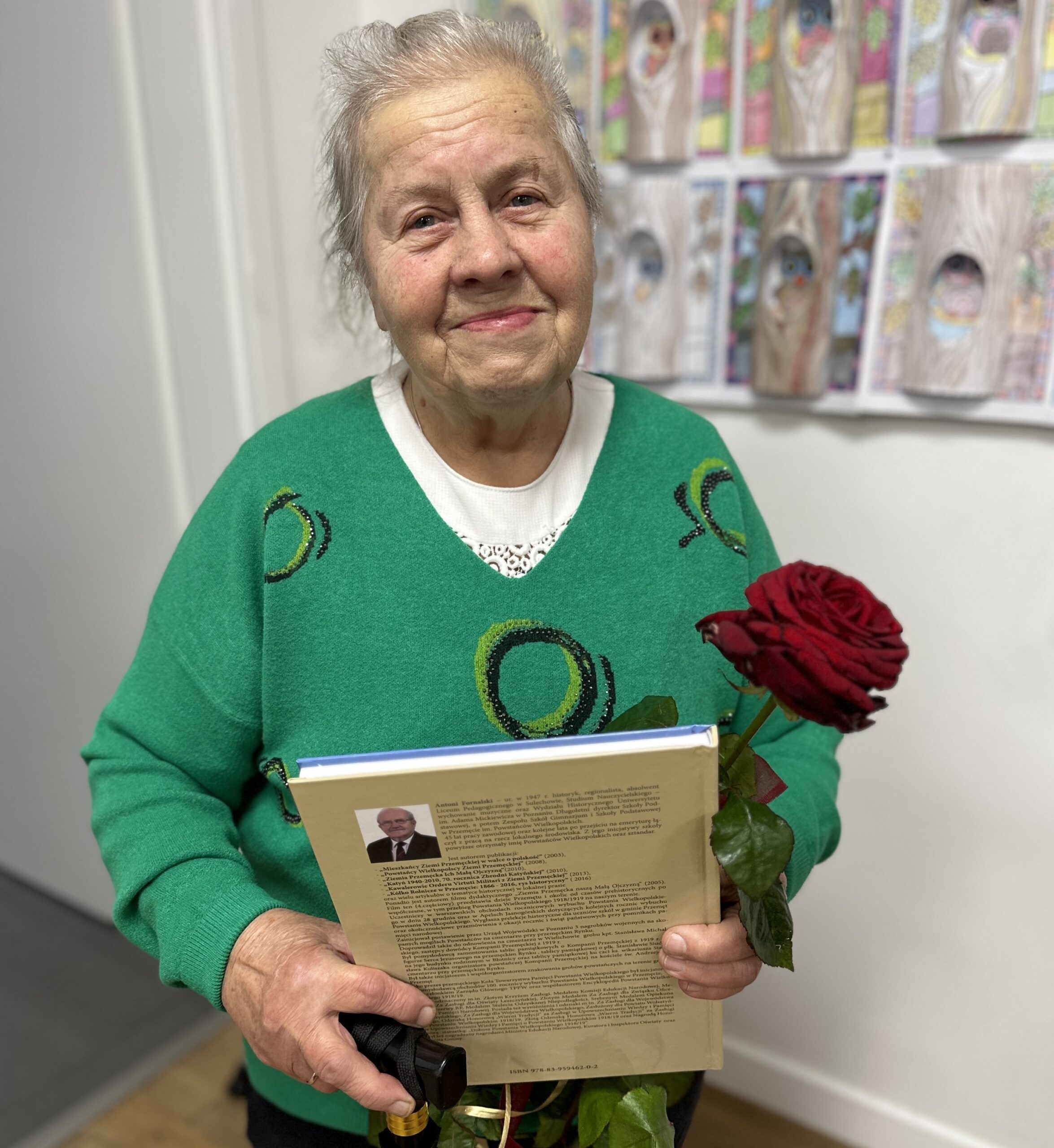 Zdjęcie przedstawia Panią Janinę, która pozuje do zdjęcia z różą i książką Antoniego Fornalskiego.