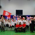Zdjęcie przedstawia wszystkich uczniów biorących udział w występach artystycznych. Uczniowie stoją na środku sali wymachując flagami Polski oraz Powstania Wielkopolskiego.