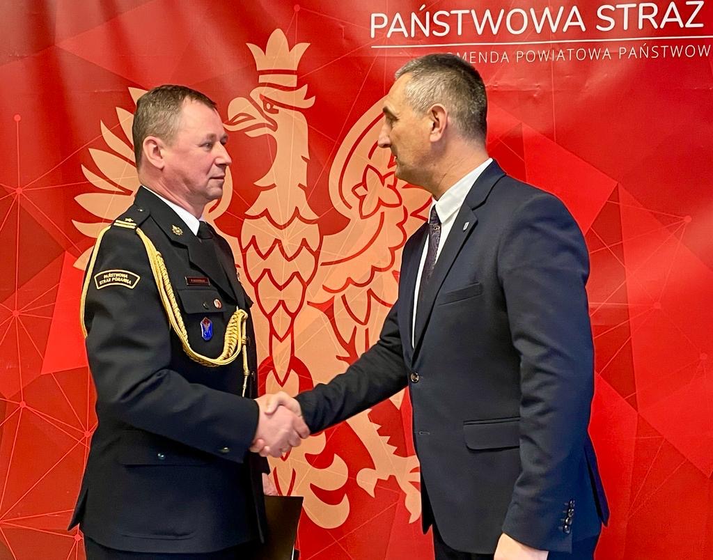 Zdjęcie przedstawia uścisk dłoni Wójta Gminy Przemęt z nowym komendantem kpt. Damianem Glapiakiem. W tle czerwona ścianka, na której widnieje wizerunek orła symbolizującego godło Polski oraz napis "Państwowa Straż Pożarna".