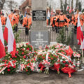 Zdjęcie przedstawia pomnik, wokół którego znajdują się wiązanki oraz flagi Polski.