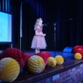 Na zdjęciu dziewczynka na scenie recytująca swój wiersz. Scena ozdobiona kolorowymi dodatkami.