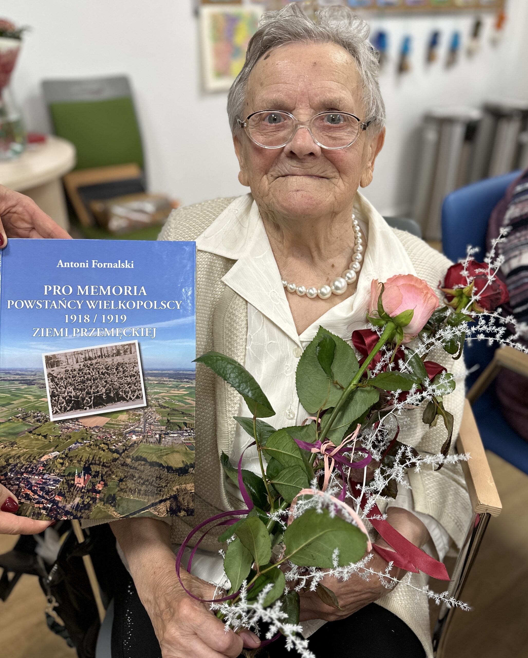 Zdjęcie przedstawia Panią Irenę Wawrzyniak trzymającą róże. Na obrazku widnieje również książka autorstwa Antoniego Fornalskiego, którą otrzymała Pani Irena.