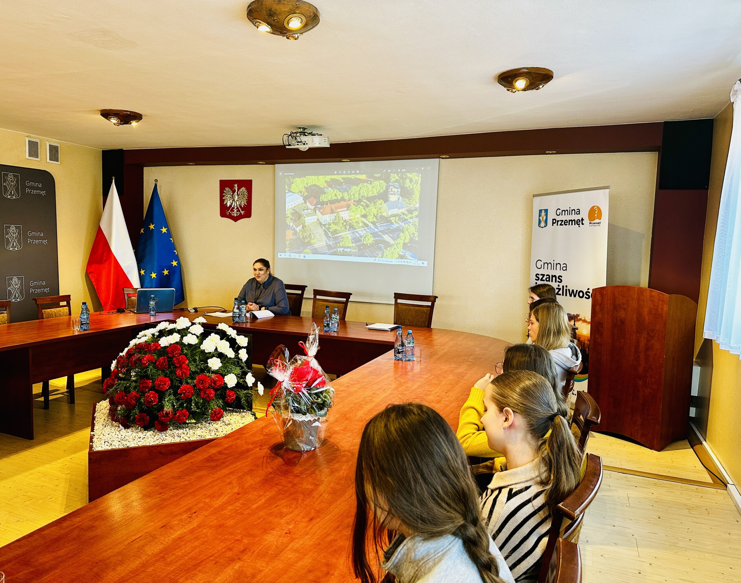 Zdjęcie przedstawia uczniów oraz Panią Martę Sobierajską siedzących na sali sesyjnej. W oddali widać zdjęcie szkoły w Mochach, które jest wyświetlane na projektorze.