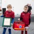 Zdjęcie przedstawia dwóch chłopców, którzy są członkami klubu SKS "Helios Bucz". Jeden z nich trzyma apteczkę pierwszej pomocy, a drugi list z gratulacjami z okazji 30-lecia klubu Helios.