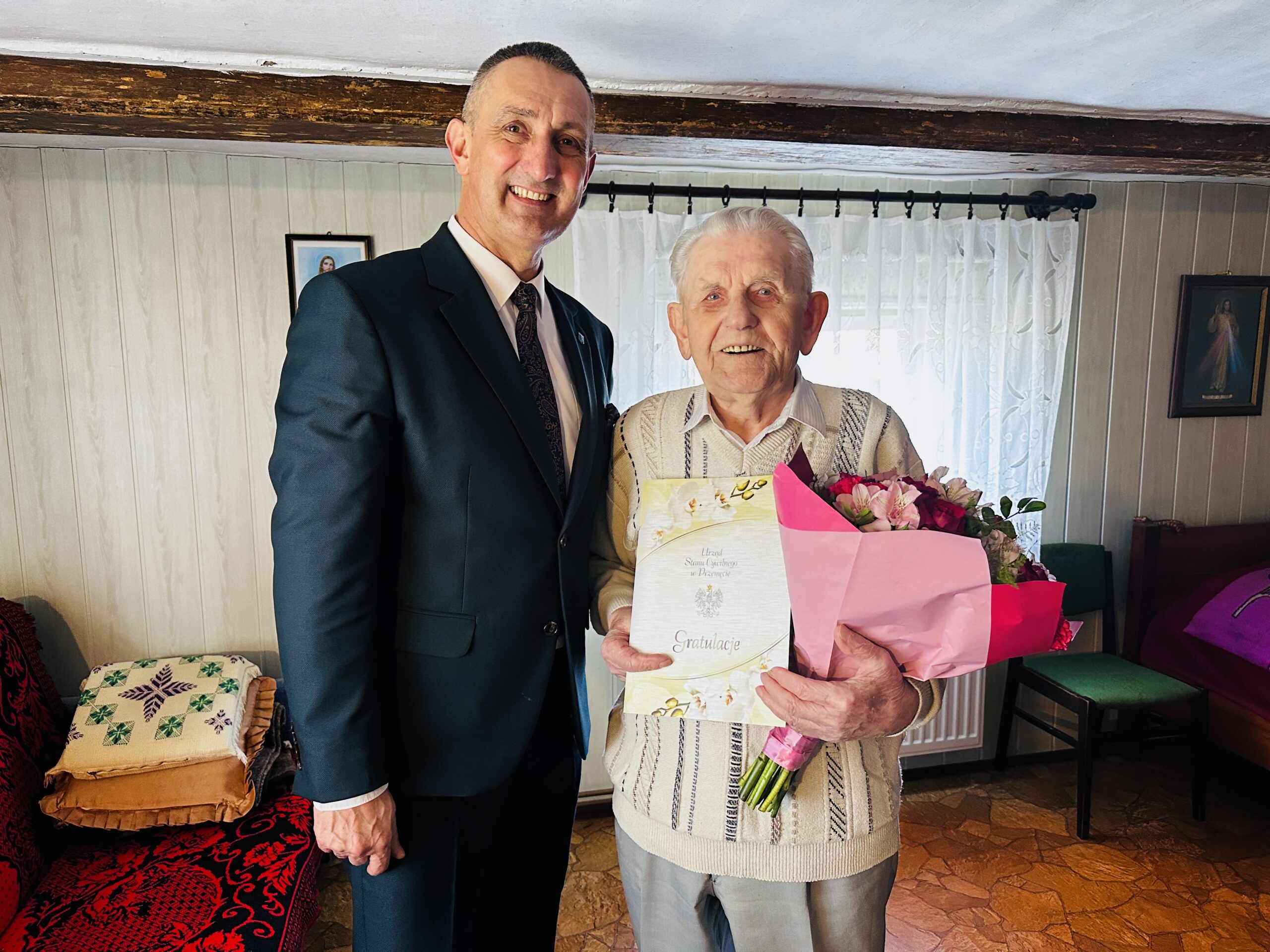 Na zdjęciu wójt i pan Zygmunt, który trzyma w rękach bukiet kwiatów oraz list gratulacyjny.