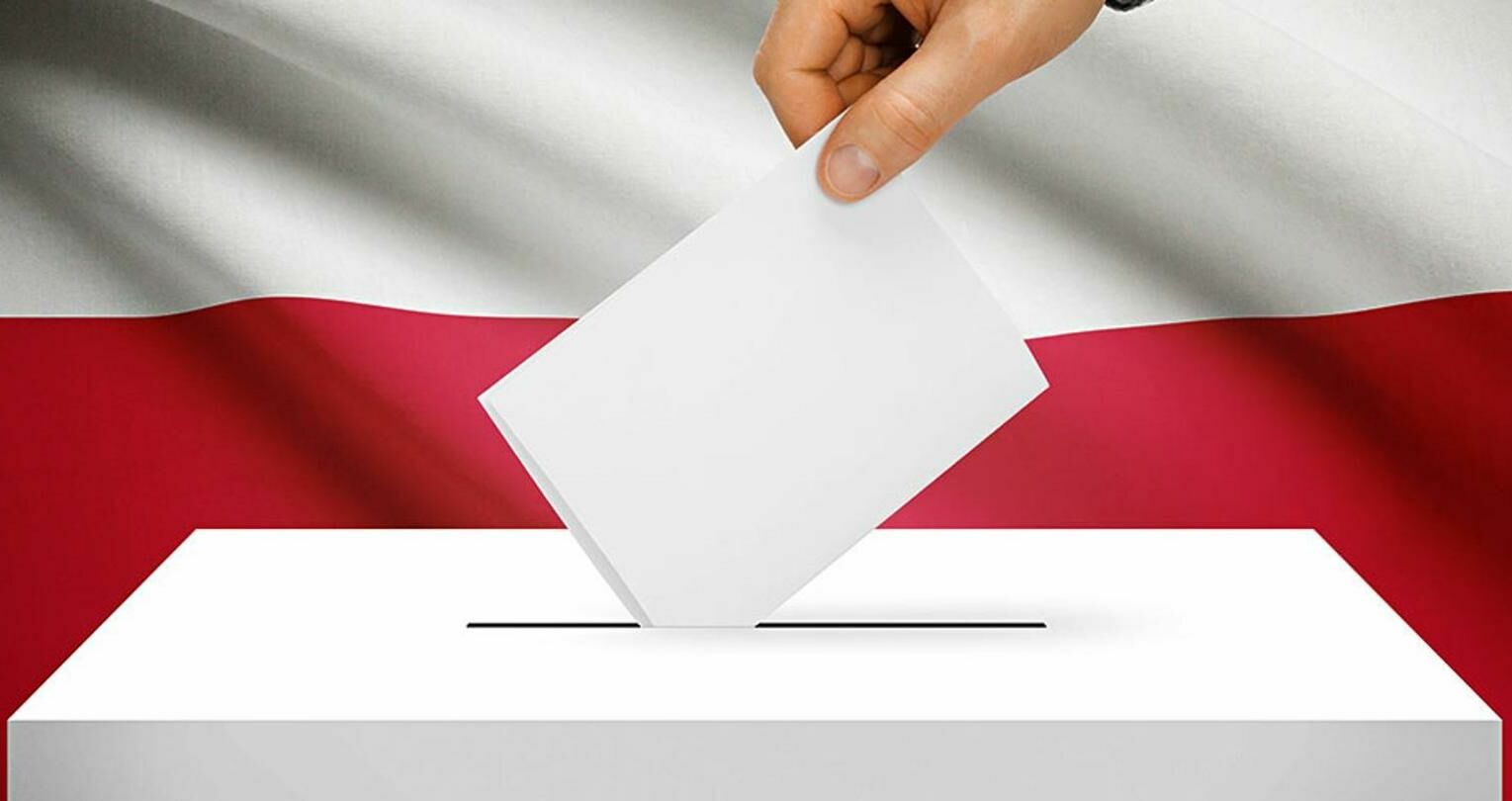 Zdjęcie przedstawia dłoń, która wrzuca kartę do głosowania do urny wyborczej. W tle flaga Polski.