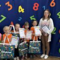 Na zdjęciu przedszkolaki z Radomierza pozują do zdjęcia z nagrodami i dyplomami na tle ścianki ozdobionej kolorowymi cyferkami oraz balonami. Na zdjęciu znajduje się też nauczycielka z przedszkola.