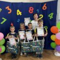 Na zdjęciu przedszkolaki ze Starkowa pozują do zdjęcia z nagrodami i dyplomami na tle ścianki ozdobionej kolorowymi cyferkami oraz balonami. Na zdjęciu znajduje się też nauczycielka z przedszkola.