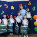 Na zdjęciu przedszkolaki z Bucza pozują do zdjęcia z nagrodami i dyplomami na tle ścianki ozdobionej kolorowymi cyferkami oraz balonami. Na zdjęciu znajduje się też nauczycielka z przedszkola.