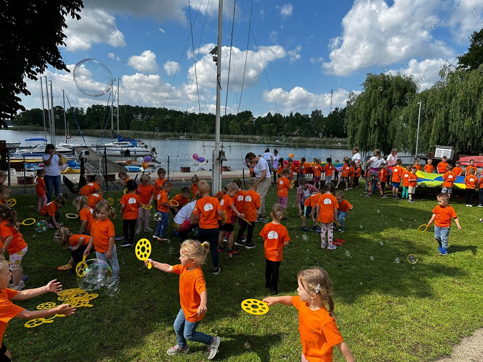 Zdjęcie przedstawia dzieci ubrane w pomarańczowe koszulki z logiem Gminy Przemęt. Zadowolone dzieci puszczają bańki. Na zdjęciu znajdują się też nauczycielki.