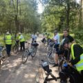 Rozproszona grupa rowerzystów stoi wraz z rowerami w lesie. Przerw regeneracyjna na łyk wody. W tle las.