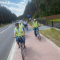 Rowerzyści poruszają się po ścieżce rowerowej wzdłuż jezdni. W tle las