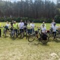 Grupa rowerzystów wraz z rowerami pozuje do grupowego zdjęcia. W tle zielone boisko oraz las.