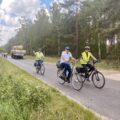 Na głównym planie trzech rowerzystów poruszających się drogą asfaltową. W tle las, traktor z przyczepą oraz inni rowerzyści. Zdjęcie zrobione z pobocza jezdni.