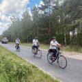 Trzech rowerzystów porusza się na rowerach drogą asfaltową. W tle las oraz przyczepa z balotami.. Zdjęcie zrobione z pobocza jezdni.