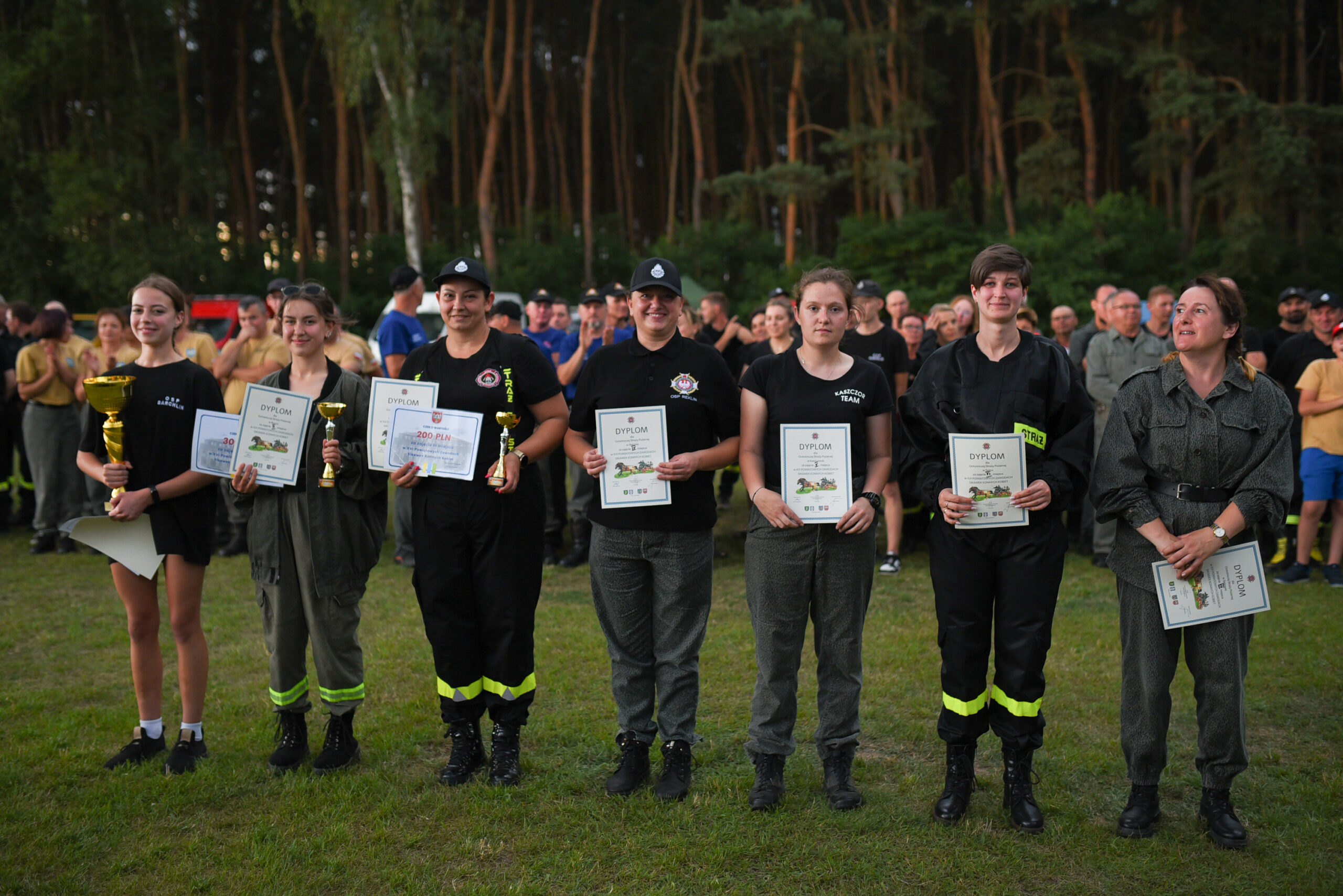 Grupowe zdjęcie kobiet, które zajęły miejsca od pierwszego do siódmego w zawodach sikawek konnych kobiet. Każda z nich trzyma w rękach dyplom, a te które zajęły miejsca od pierwszego do trzeciego trzymają także puchary i czeki.