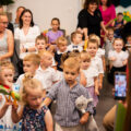 Na zdjęciu dzieci zmierzające w głąb sali, w której odbywa się uroczyste zakończenie roku. W sali siedzą także rodzice dzieci.