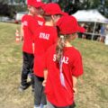 Zdjęcie przedstawia dzieci w czerwonych bluzkach z napisem "straż" i czerwonych czapkach z daszkiem. Dzieci stoją odwrócone tyłem do aparatu,