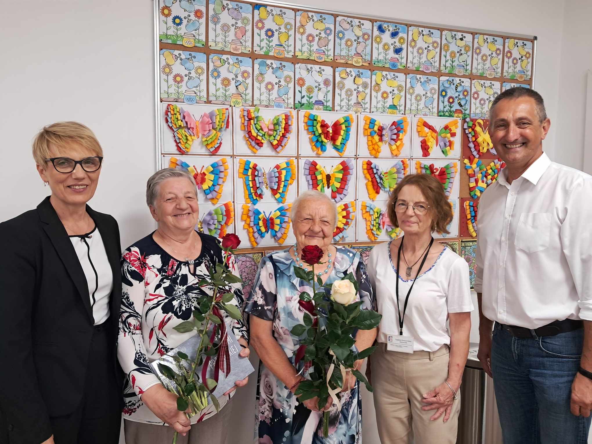 Na zdjęciu wójt, dyrektor GOPSu Barbara Ścigała, kierownik DDPS+ Izabela Kwaczyńska oraz dwie jubilatki, które trzymają w rękach róże. Zdjęcie zrobione na tle prac artystycznych.