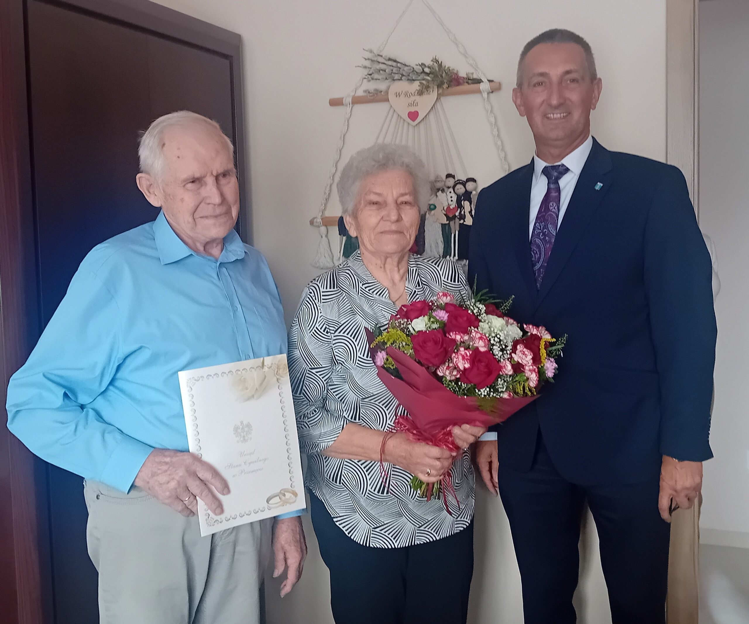 Zdjęcie przedstawia państwa Marię i Leona Kokornarczyk wraz z wójtem Gminy Przemęt Januszem Frąckowiakiem. Pani Maria trzyma w rękach kwiaty, natomiast Pan Leon trzyma w rękach teczkę z listem gratulacyjnym z okazji 60 rocznicy ślubu.