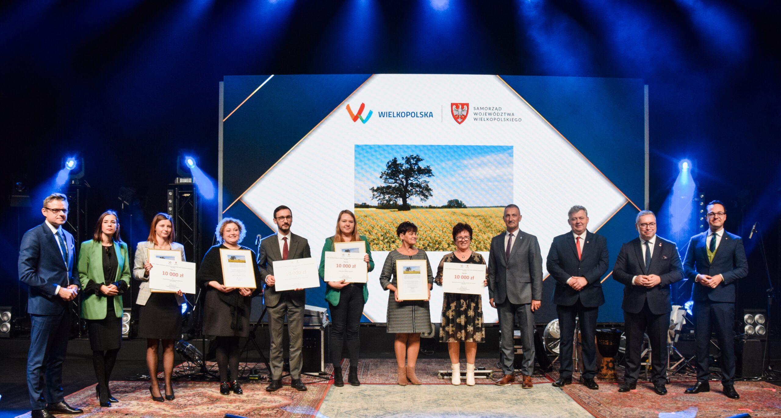 Wspólne zdjęcie na scenie. Na zdjęciu pozują Pan Jacek Bogusławski, wójt Janusz Frąckowiak, starosta Jacek Skrobisz oraz przedstawiciele sołectw, szkół i stowarzyszenia. Nagrodzeni trzymają nagrody.