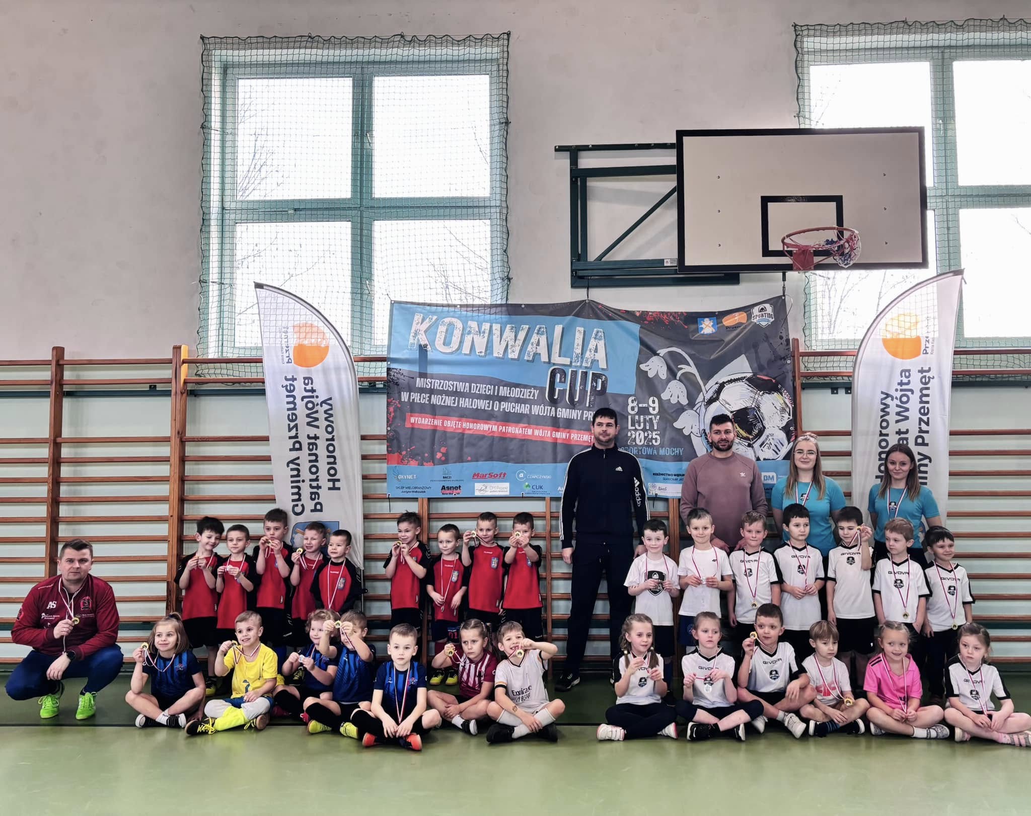 Grupowe zdjęcie drużyn z danej kategorii wiekowej. Zawodnicy mają założone medale. Na zdjęciu także ich trenerzy. Na fotografii znajduje się baner Konwalia Cup oraz dwa windery z honorowym patronatem wójta gminy Przemęt.