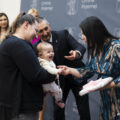 Na zdjęciu przewodnicząca wręcza mamie dziecka upominek. Dziecko jest uśmiechnięte. Na fotografii znajduje się również wójt, który wskazuje palcem na upominek.
