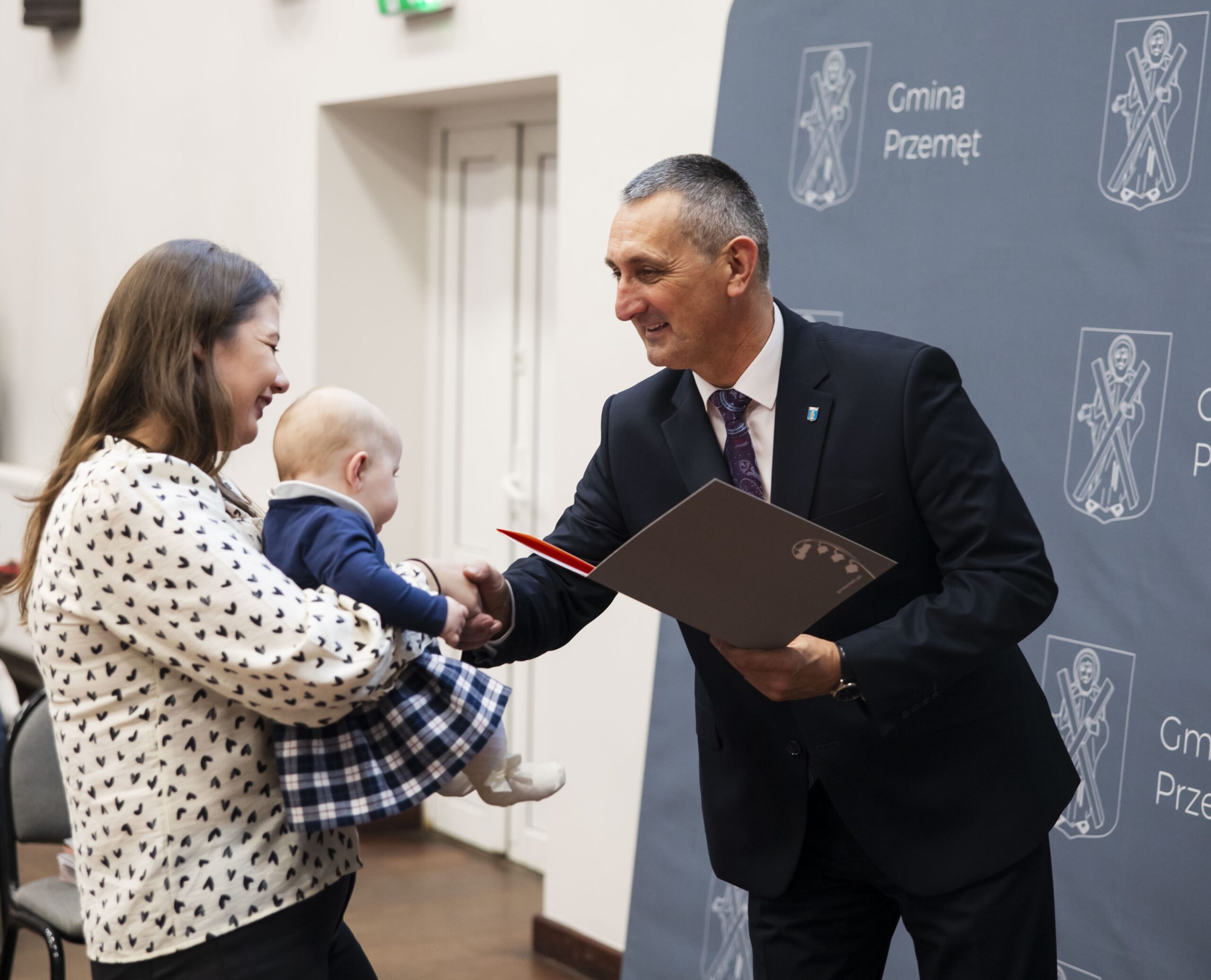 Na zdjęciu wójt składa gratulacje oraz wręcza list gratulacyjny matce, która trzyma dziecko na rękach.