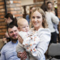 Na zdjęciu uśmiechnięta mama wraz z dzieckiem, które trzyma na rękach. W tle ojciec dziecka.