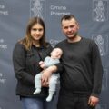 Na zdjęciu rodzice wraz z dzieckiem pozują na dużej szarej ściance, która zawiera herb gminy Przemęt oraz napisy "Gmina Przemęt".