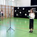 Na fotografii dziewczyna, która przemawia podczas zakończenia roku szkolnego na sali gimnastycznej.