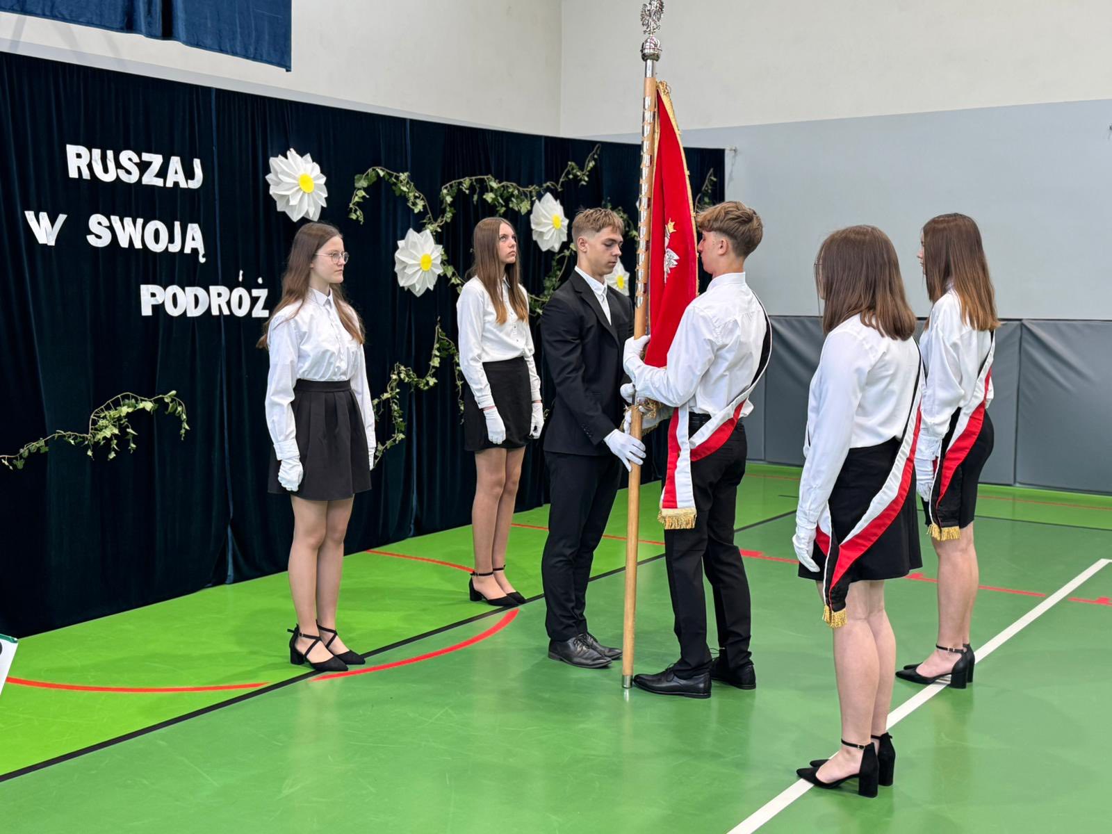 Na fotografii uchwycono moment przekazania sztandaru. Na zdjęciu szóstka uczniów. W tle ścianka z napisem "Ruszaj w swoją podróż".