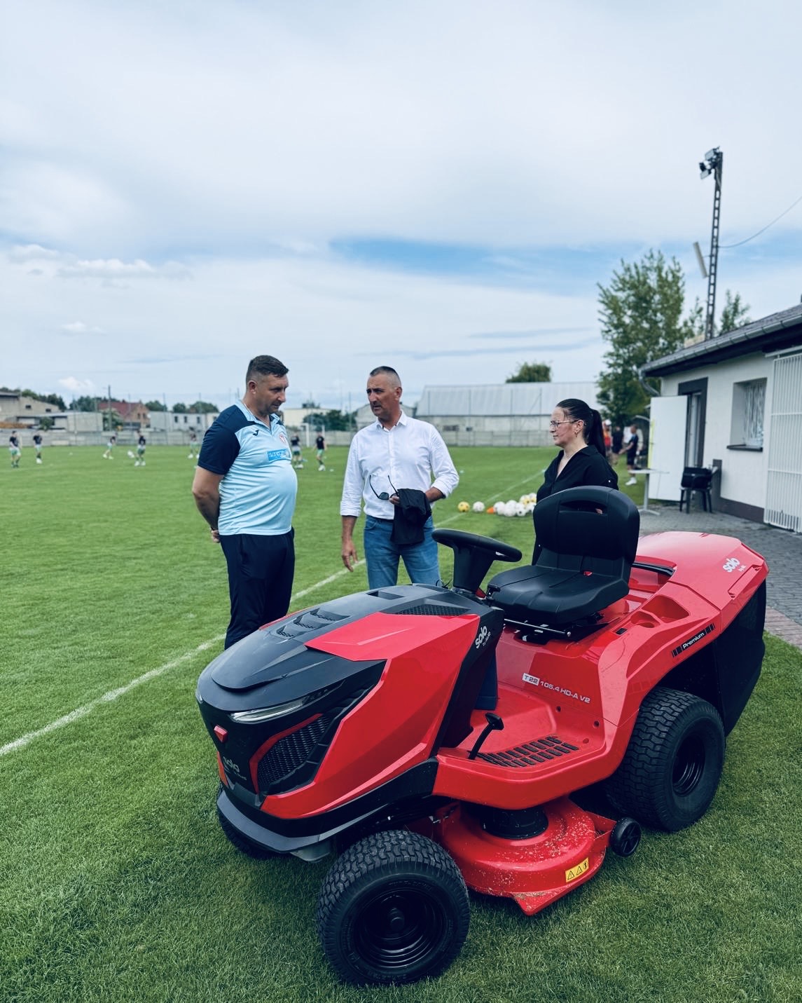 Na fotografii wójt gminy Janusz Frąckowiak, radna gminy Kinga Piędel oraz prezes klubu sportowego Sokół Kaszczor. Wyżej wymienieni rozmawiają. Na zdjęciu także nowy czerwony traktor ogrodowy.