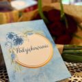 Na fotografii widoczna niebieska kartka z grafiką niebieskiego kwiatka i złotymi elementami. Na środku kartki widnieje napis: "Podziękowanie". Kartka ma format A5.