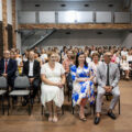 Zdjęcie przedstawia osoby siedzące na widowni. Na fotografii m.in. wójt, przewodnicząca rady oraz kierownik oświaty, a także dyrektorzy szkoły i zaproszeni uczniowie wraz z rodzicami.
