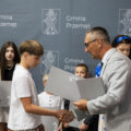 Na fotografii wójt wręcza nagrodę jednemu z nagrodzonych uczniów. W tle inni uczniowie, przewodnicząca rady oraz kierownik oświaty.