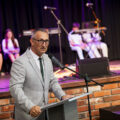 Zdjęcie przedstawia wójta, który przemawia zza mównicy podczas uroczystości. W tle scena.
