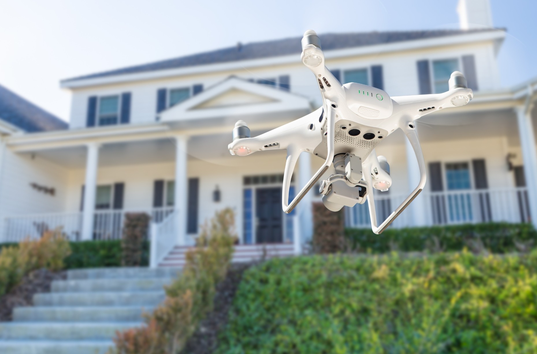 To zdjęcie przedstawia drona unoszącego się nad terenem, z wyraźnym widokiem na przednią część domu. Jest to klasyczny quadcopter, który posiada kamerę zamocowaną na dolnej części konstrukcji. Dron znajduje się w powietrzu.