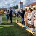 Zdjęcie ukazuje moment wręczenia dożynkowego daru przez delegację mieszkańców. Kobieta i mężczyzna przekazują księdzu bukiet zbóż i kwiatów – tradycyjny symbol wdzięczności za plony. W tle widać uczestników wydarzenia ubranych odświętnie, stojących w rzędach na trawie przy ławkach ustawionych w plenerze.