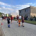 Na zdjęciu grupa dzieci i młodzieży w strojach sportowych bierze udział w grze w piłkę nożną na ulicy. Dzieci ubrane są w koszulki z numerami, a wokół ustawione są czerwone pachołki. Po bokach stoją dorośli i kibice.