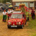 Na zdjęciu widoczny jest kolorowy korowód dożynkowy prowadzony przez czerwonego „malucha” (Fiat 126p), udekorowanego kwiatami, zbożem i napisem "Sycewice". Za pojazdem ciągnięta jest platforma z wieńcem dożynkowym, z którego wychodzą czerwone wstęgi trzymane przez uczestników w tradycyjnych strojach ludowych. W tle inni ludzie.