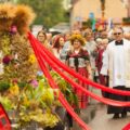 Na fotografii widoczna jest część korowodu dożynkowego. W centrum znajduje się wieniec dożynkowy ozdobiony kwiatami, wstążkami i kłosami zbóż. Obok idą uczestnicy ubrani w stroje regionalne, w tym kobieta z wiankiem z kwiatów i ziół oraz ksiądz w albie i stułą, który towarzyszy procesji. W tle widać barwną grupę mieszkańców i gości.