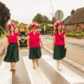 Na zdjęciu trzy kobiety ubrane w jednolite stroje – różowe bluzki, ciemnozielone spódnice i efektowne wianki z kwiatów i zbóż – maszerują dumnie ulicą w ramach pochodu dożynkowego. W tle widoczny jest wieniec niesiony na platformie oraz pozostali uczestnicy korowodu.