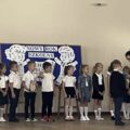 Na zdjęciu znajdują się uczniowie najmłodszych klas, ustawieni w rzędzie podczas oficjalnej części rozpoczęcia roku. Z prawej strony stoi pani dyrektor, która pasuje jednego z uczniów dużym drewnianym ołówkiem i nauczycielka, która wręcza uczniowi dyplom. W tle widać tablicę dekoracyjną z napisem „Nowy Rok Szkolny".