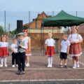 Na zdjęciu dzieci stoją w rzędzie na tle wiejskiego placu, podczas występu. Ubrane są w stroje ludowe – dziewczynki mają białe bluzki i czerwone spódniczki z kwiatowym wzorem, a chłopcy – białe koszule, jeden chłopiec trzyma mikrofon i ma kaszkiet. W tle – scena z zielonym namiotem i głośnikami, a za ogrodzeniem – ceglany mur i wiejski dom.